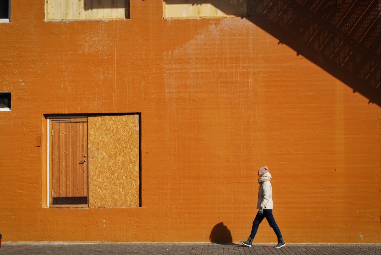 A person walks along a vibrant orange wall, casting a distinct shadow in the city.